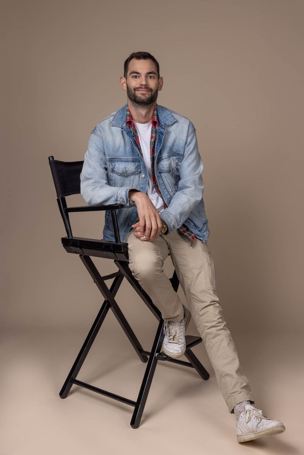 Smiling man in denim jacket and khakis leaning on director's chair against beige background.