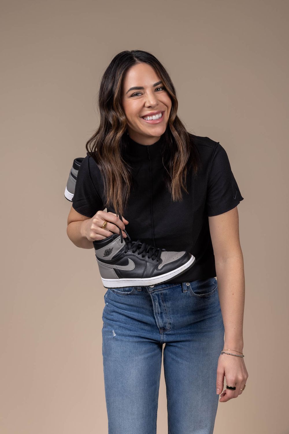 Smiling woman in black turtleneck and jeans holding Air Jordan sneaker against beige background.