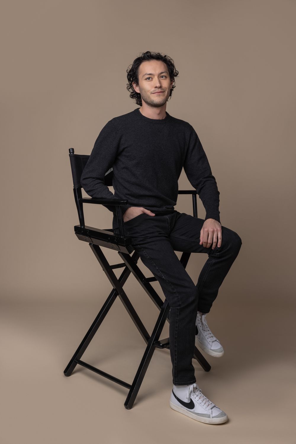 Relaxed man in all-black outfit sits in director's chair wearing white Nike sneakers.
