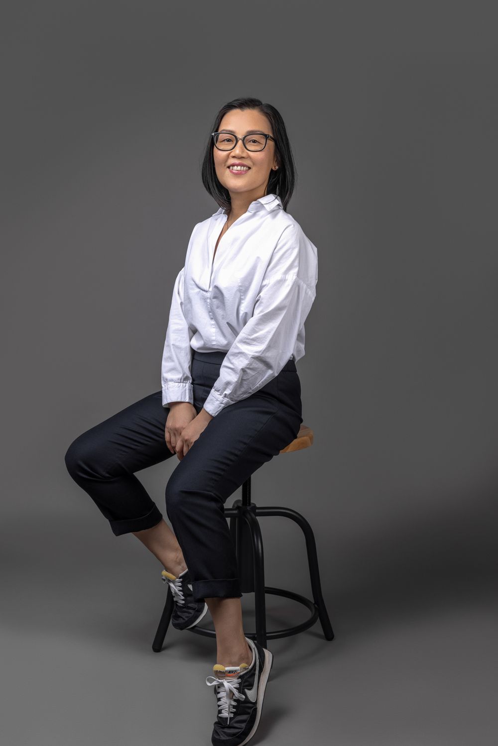 Asian woman with glasses in white shirt and black pants smiling while seated on stool against gray background.