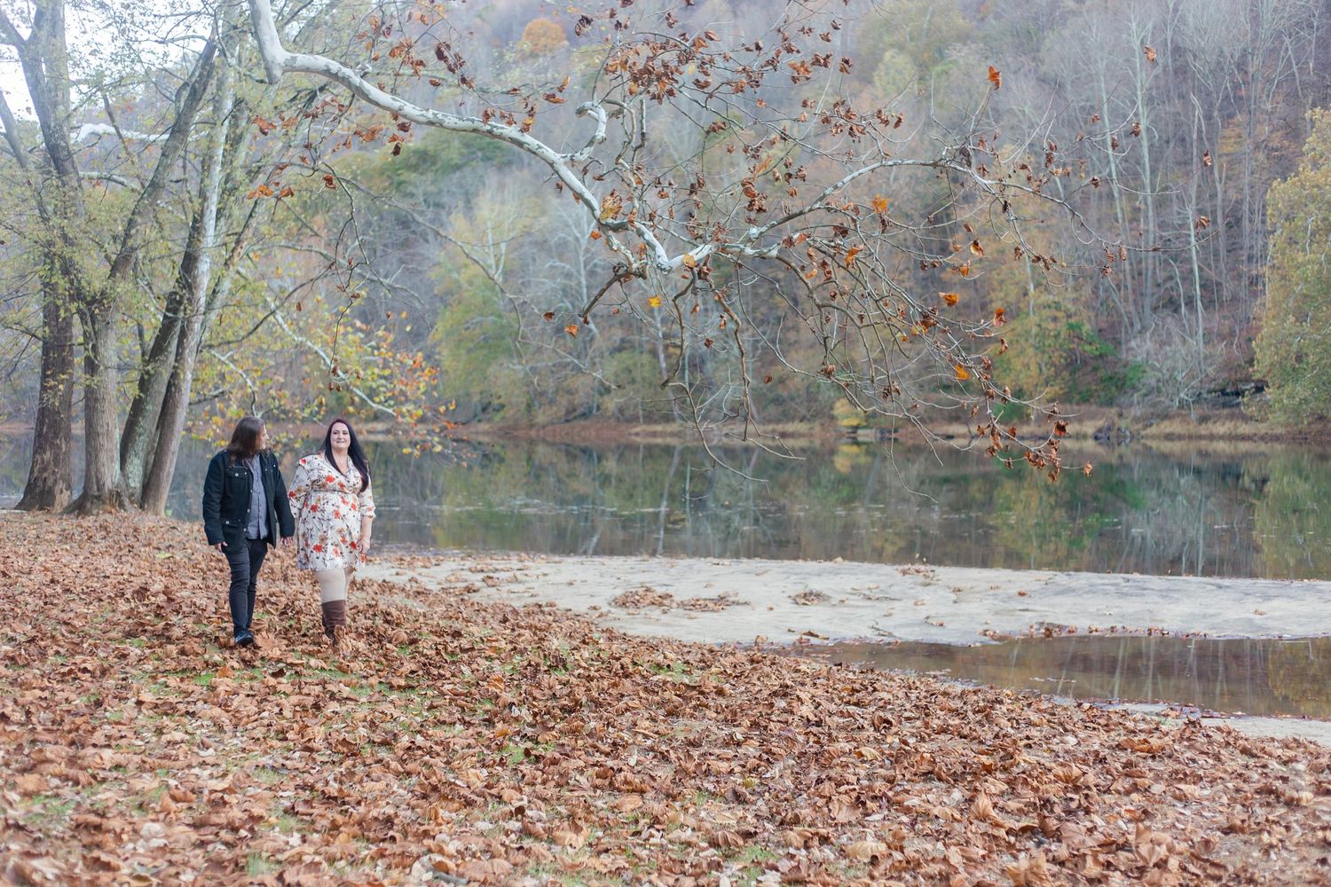 A couple walks along a leaf-covered path by a serene lake surrounded by autumn trees.