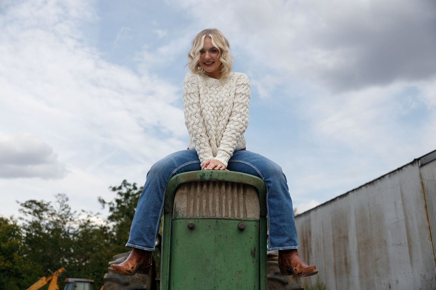 Senior lady sits on hood of green tractor for her high school senior portraits.