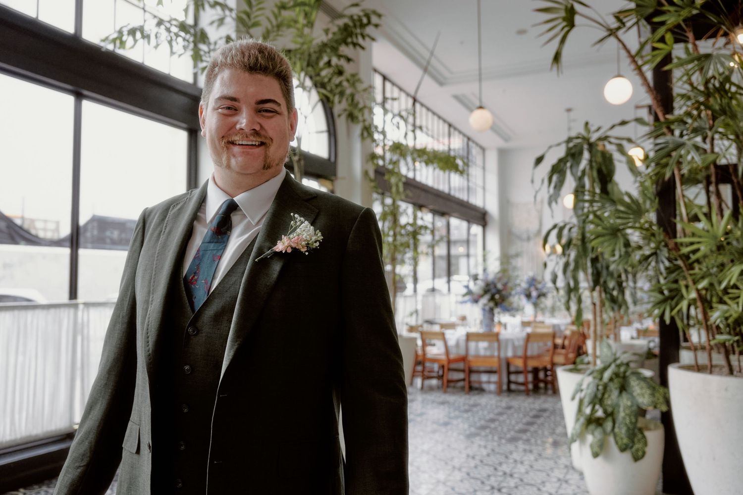 A groom in a dark suit and tie stands in an elegant venue with tropical plants and natural lighting.