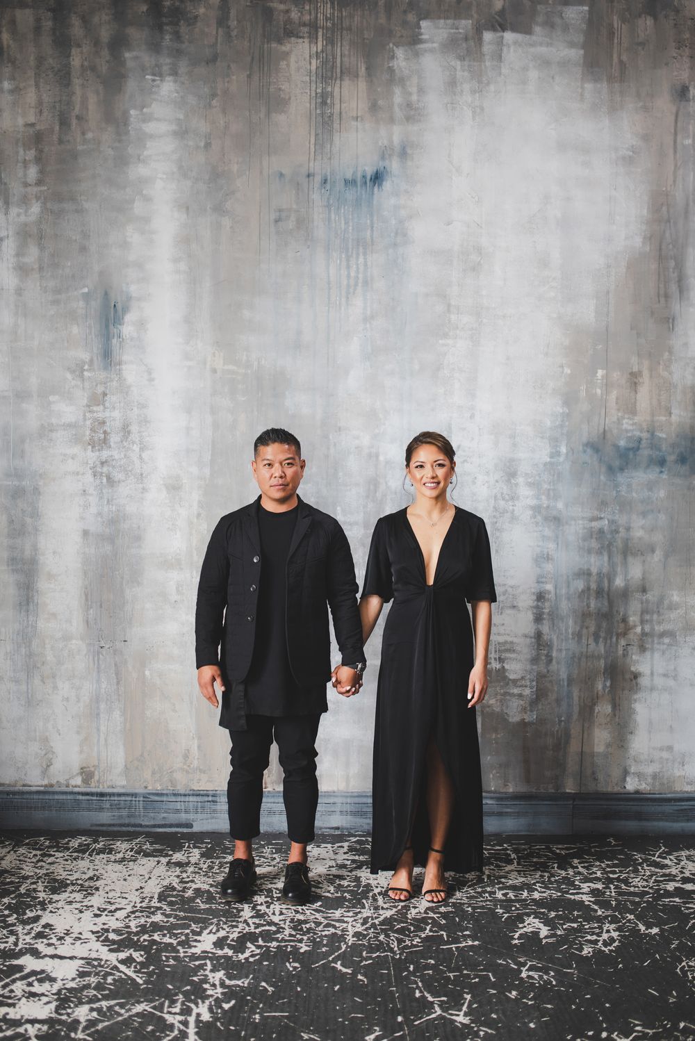 Couple in black attire holds hands while standing against textured grey wall.