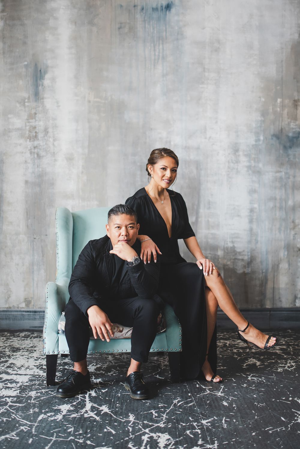 A couple poses dramatically on a turquoise chair against an industrial concrete backdrop.