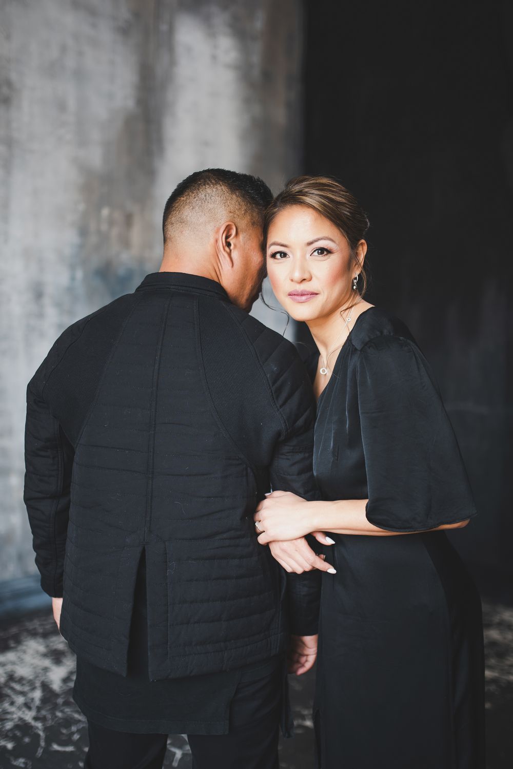 A couple in black attire poses intimately against an industrial concrete wall.
