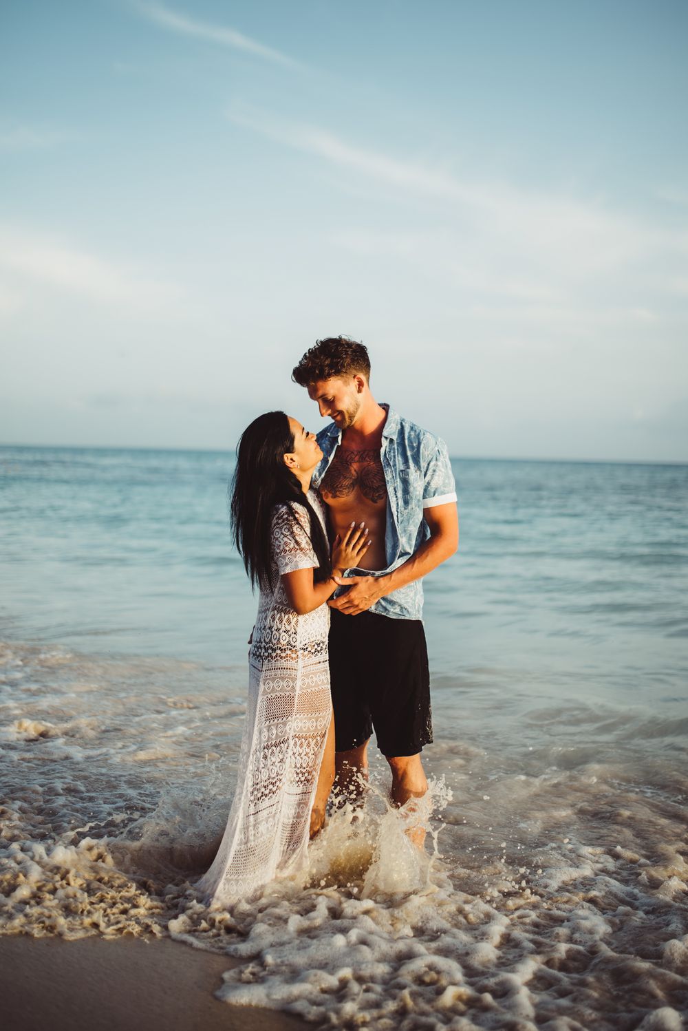 A couple stands in ocean waves wearing beach attire during a golden hour photoshoot.