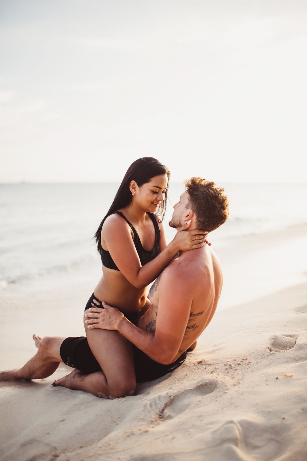 A romantic couple embraces while sitting on a sandy beach at sunset.