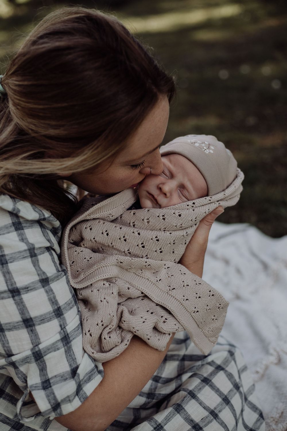 A tender moment captured as someone leans down to kiss a swaddled baby outdoors.