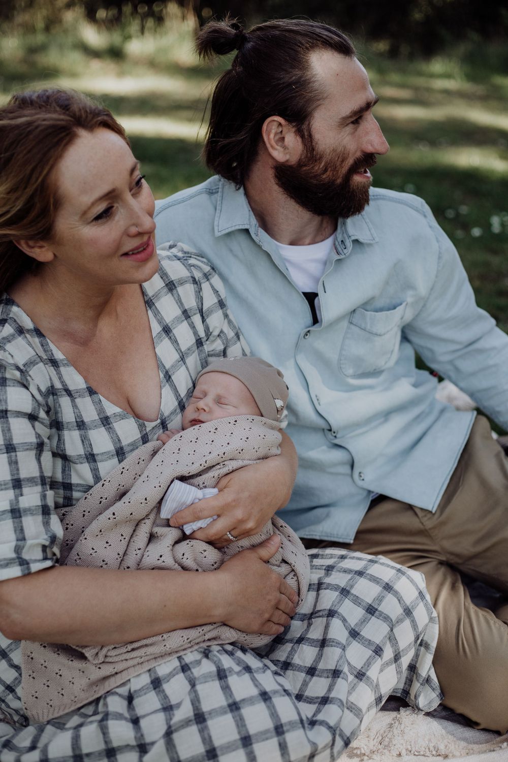 A couple in plaid shirts sit outdoors while holding their newborn baby wrapped in a patterned blanket.