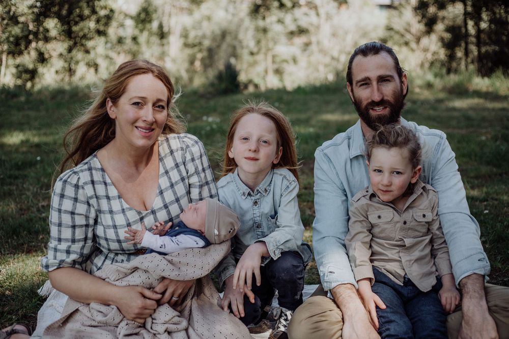 A family sits together on a blanket outdoors during a casual portrait session in dappled sunlight.