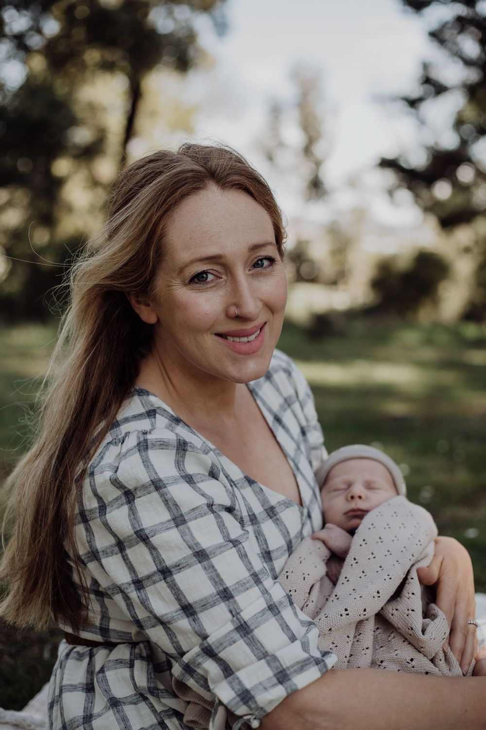 A mother in plaid sits outdoors on a sunny day while holding her sleeping newborn.