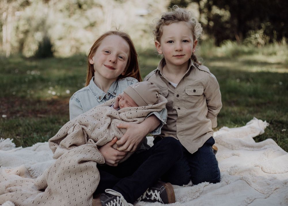 Siblings sit together on a picnic blanket outdoors sharing a tender moment.