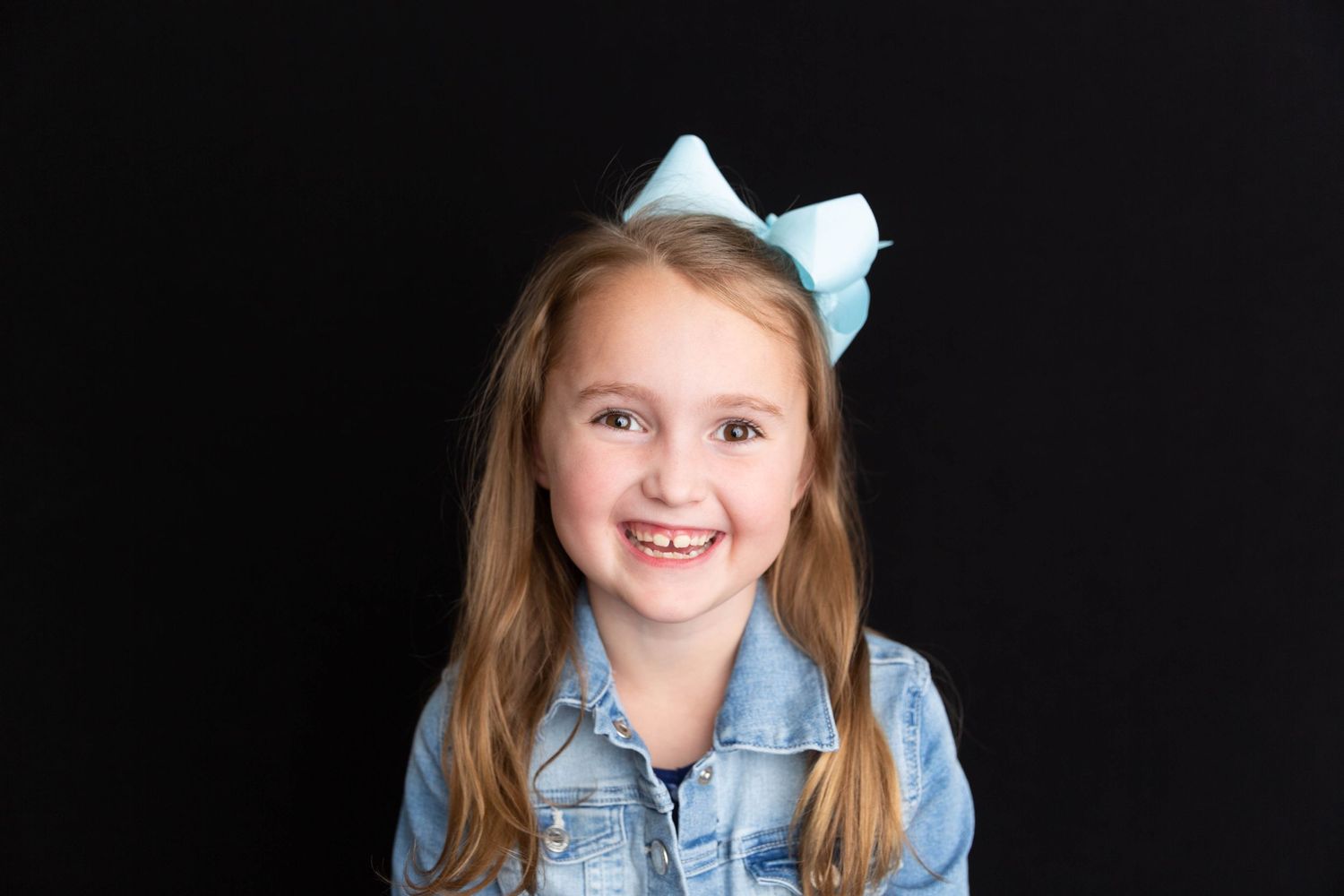 Child wearing denim jacket and blue bow against black background.