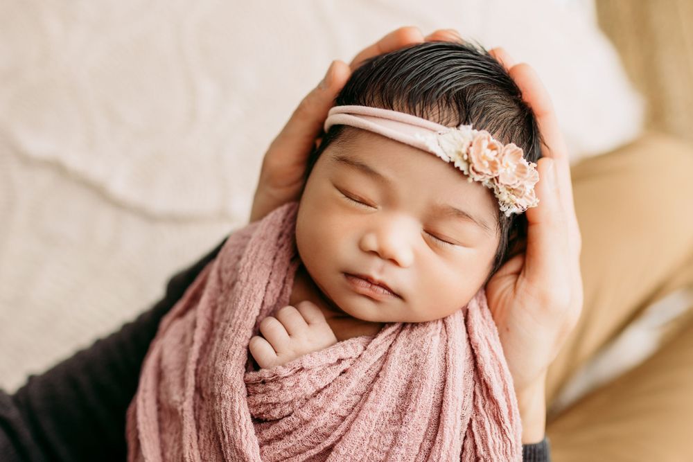 Sleeping newborn with floral headband wrapped in pink against neutral background.