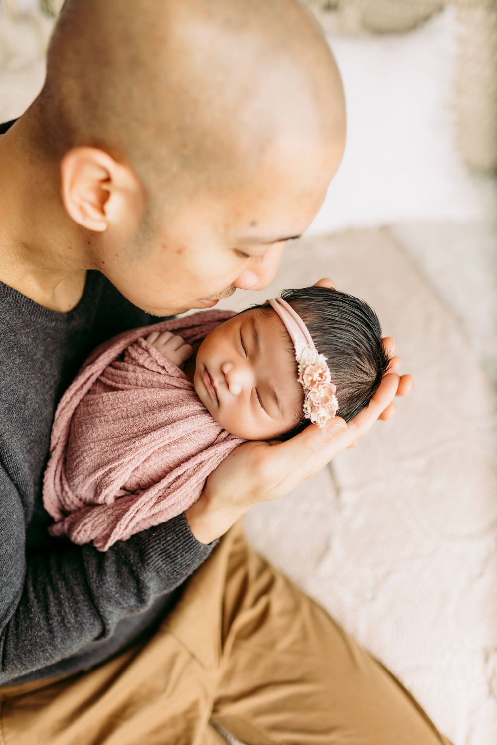 Father tenderly holding sleeping newborn wearing pink headband and wrap.