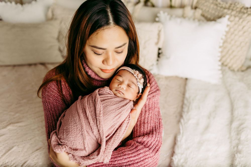 Mother in pink sweater holding sleeping newborn wrapped in soft pink blanket.