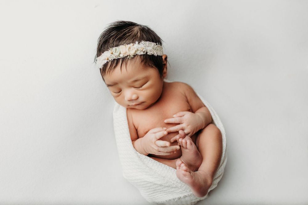 Sleeping newborn with white headband wrapped in white fabric against light background.