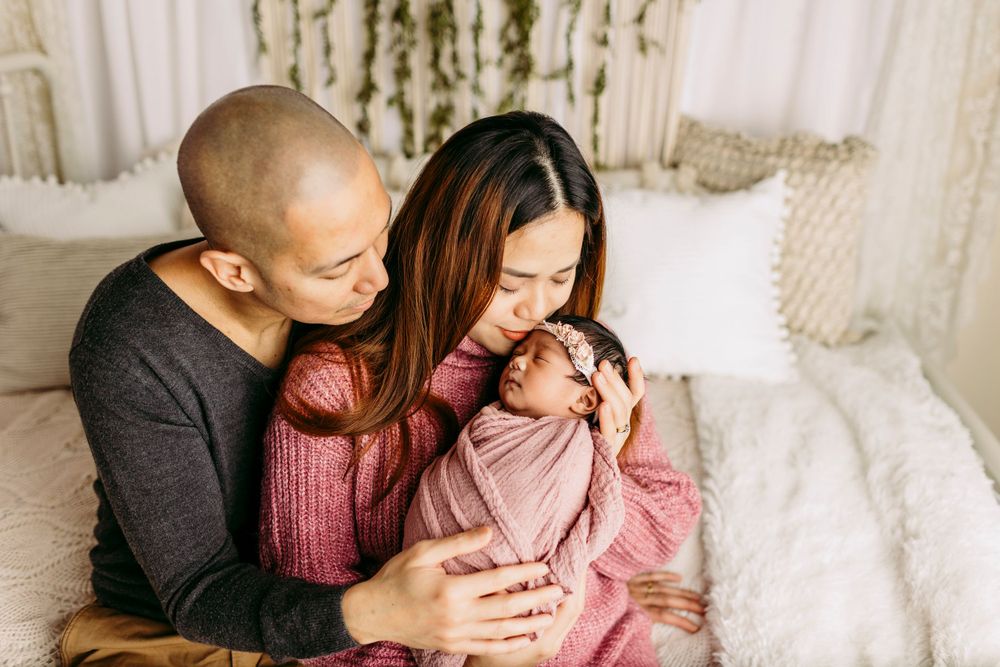Parents in casual attire holding their newborn wrapped in pink on white couch.