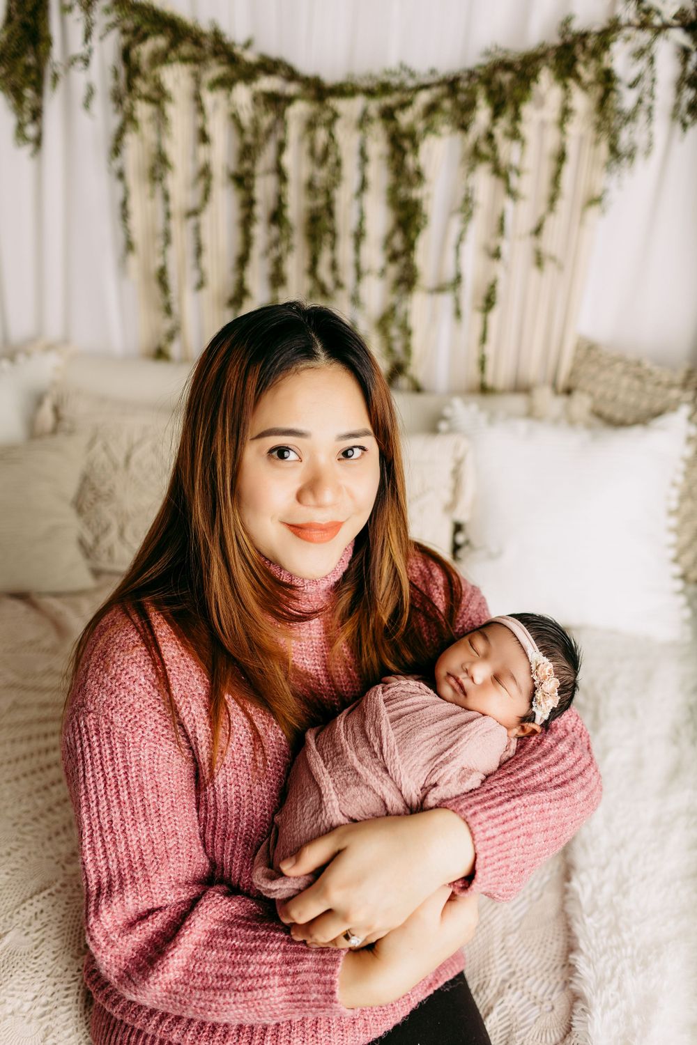 Mother in pink sweater holding sleeping newborn with evergreen garland backdrop.