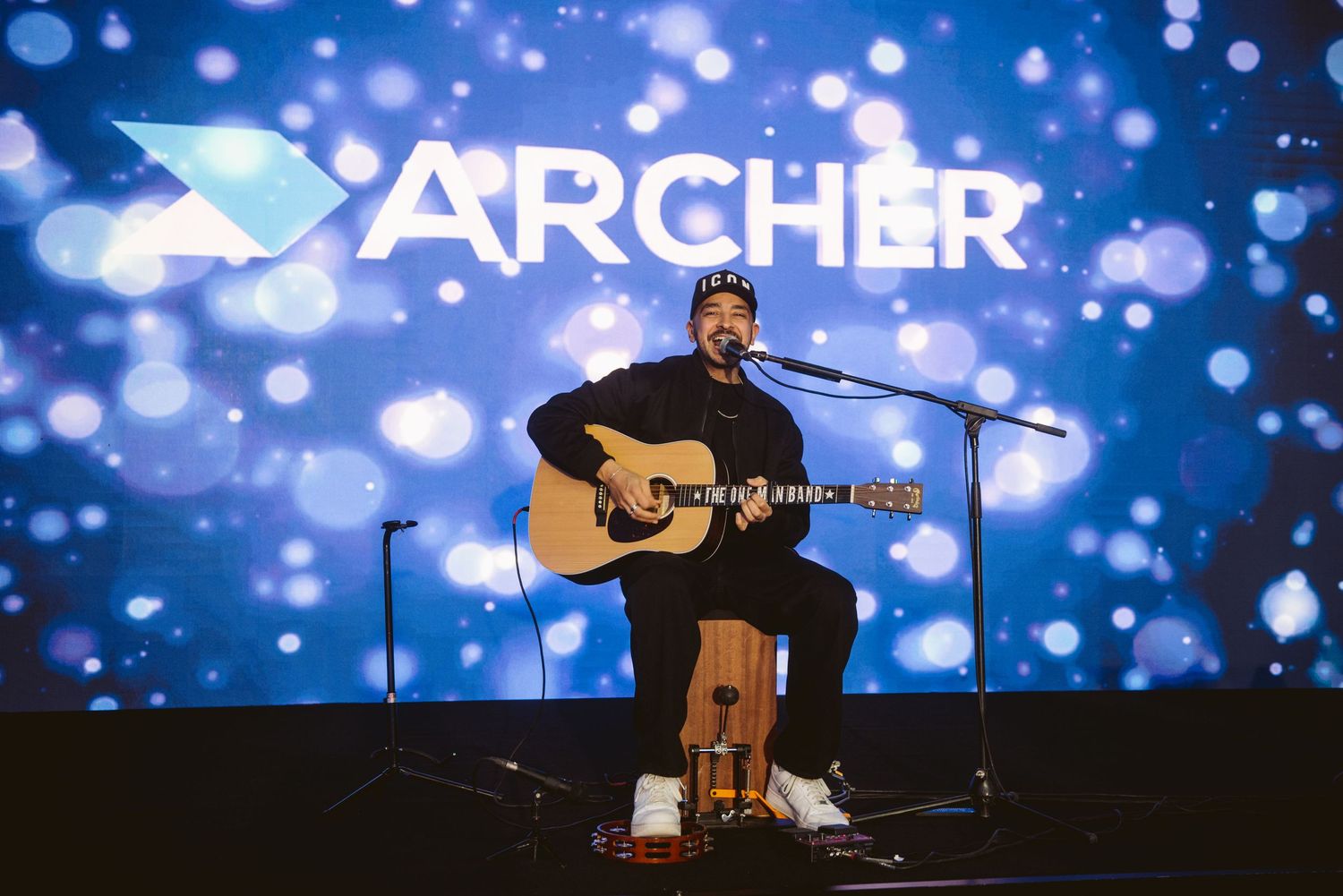 A musician performs on acoustic guitar on stage with blue glowing Archer text and bokeh lights in the background.
