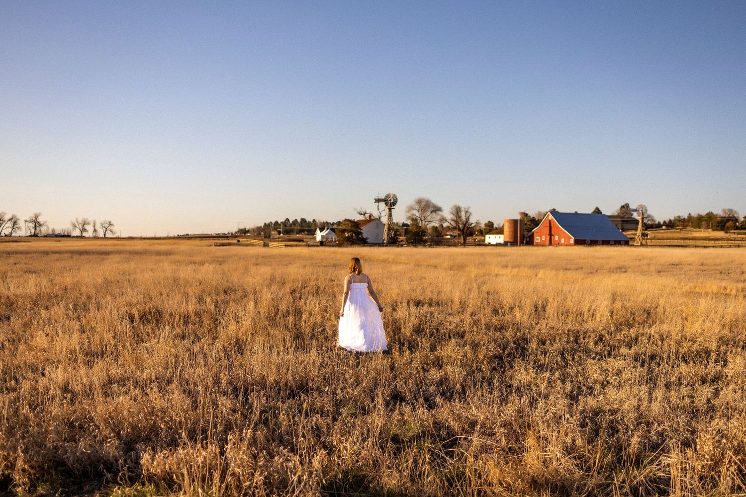 woman in long white dress walking in a field of long grass, red barn in distance and blue skies