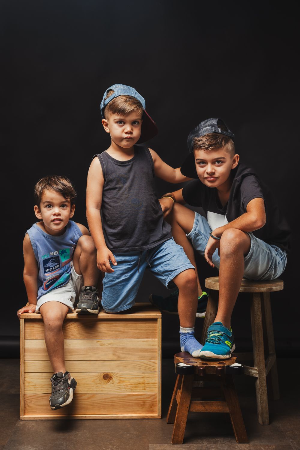 Children sitting casually on wooden furniture against black background in studio setting.