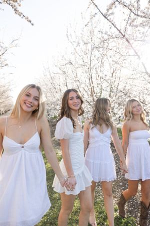 Four young women in white dresses hold hands among blooming spring trees.
