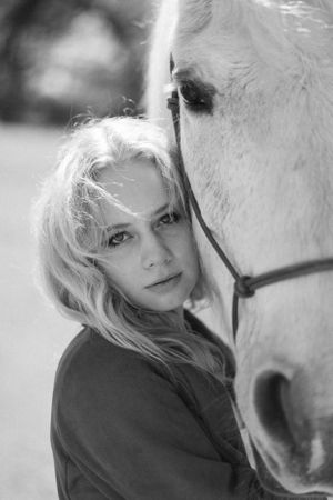 Black and white portrait of a blonde woman leaning her face against a white horse.