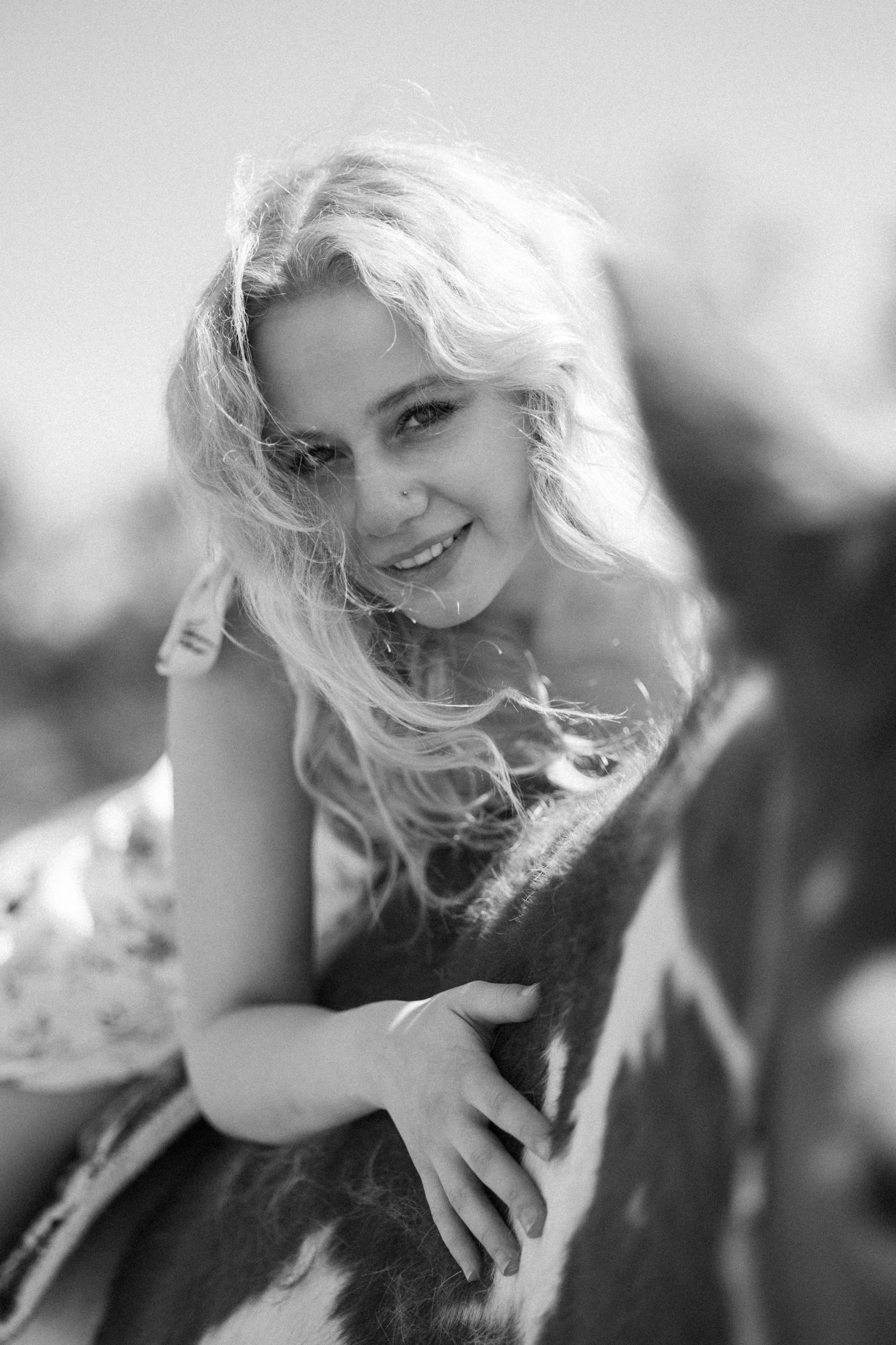 Black and white photo of a smiling blonde woman leaning over a horse in an outdoor setting.