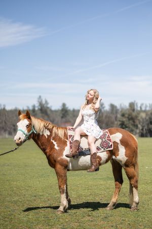 A blonde woman in a floral dress rides a paint horse in a sunny green meadow.