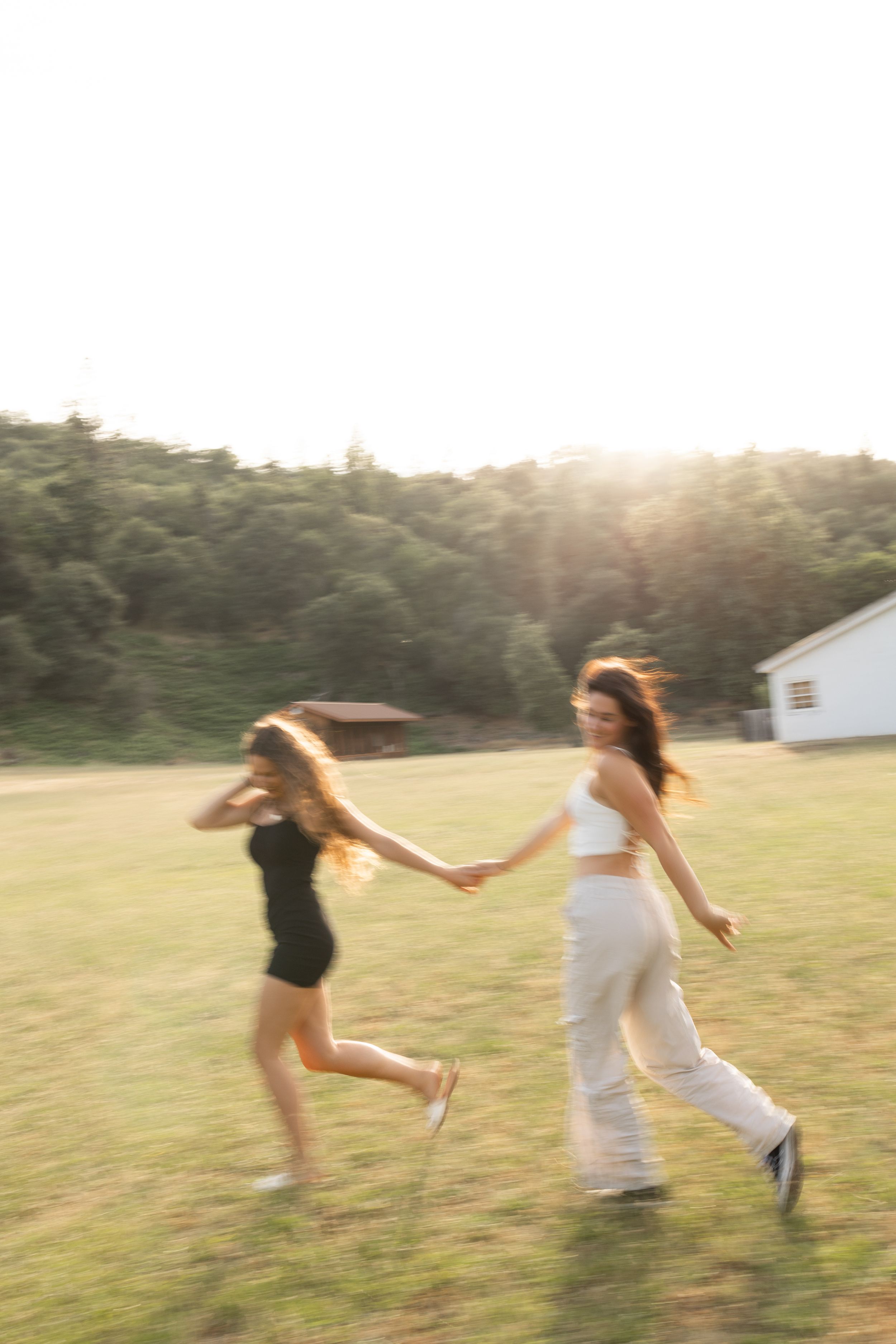 Two young women run hand in hand across a grassy field in golden sunset light.