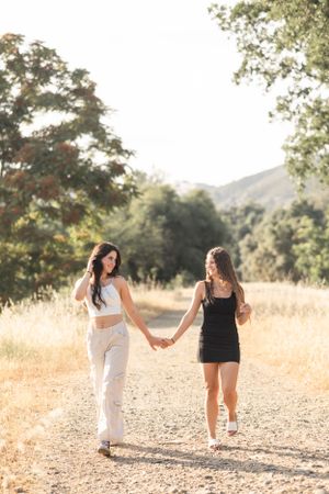 Two young women hold hands walking down a sunny dirt path surrounded by trees.