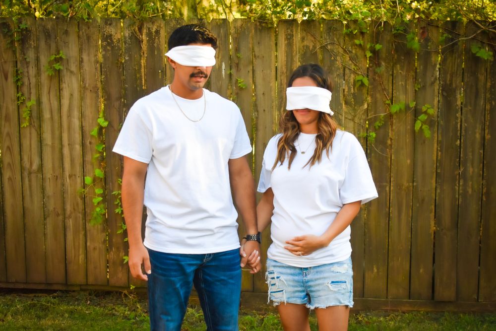 A couple in matching white shirts and blindfolds stand together in front of a wooden fence.