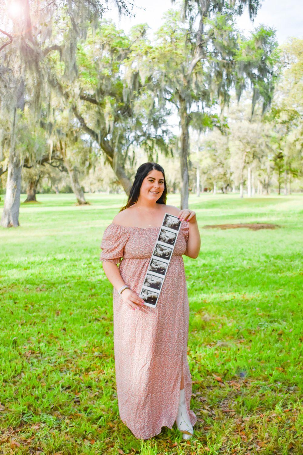 Person in pink maxi dress holding ultrasound photos in park setting.