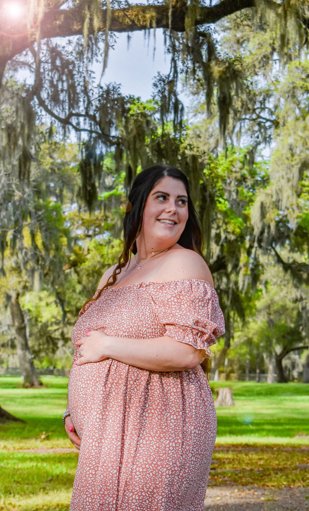 Smiling person in pink off-shoulder dress poses in park with Spanish moss trees.
