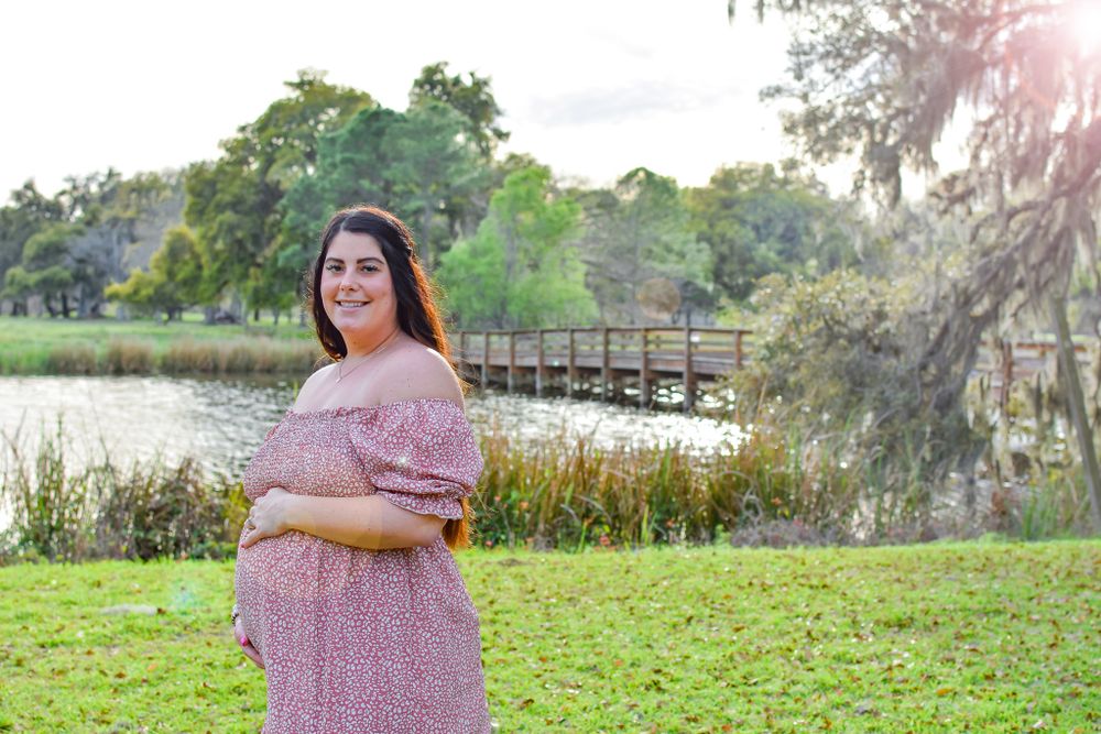 Expectant mother in pink dress standing by lake with wooden bridge in background.