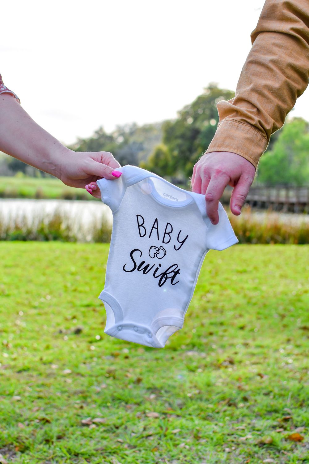 Hands holding up white baby onesie with 'Baby Swift' text in outdoor setting.
