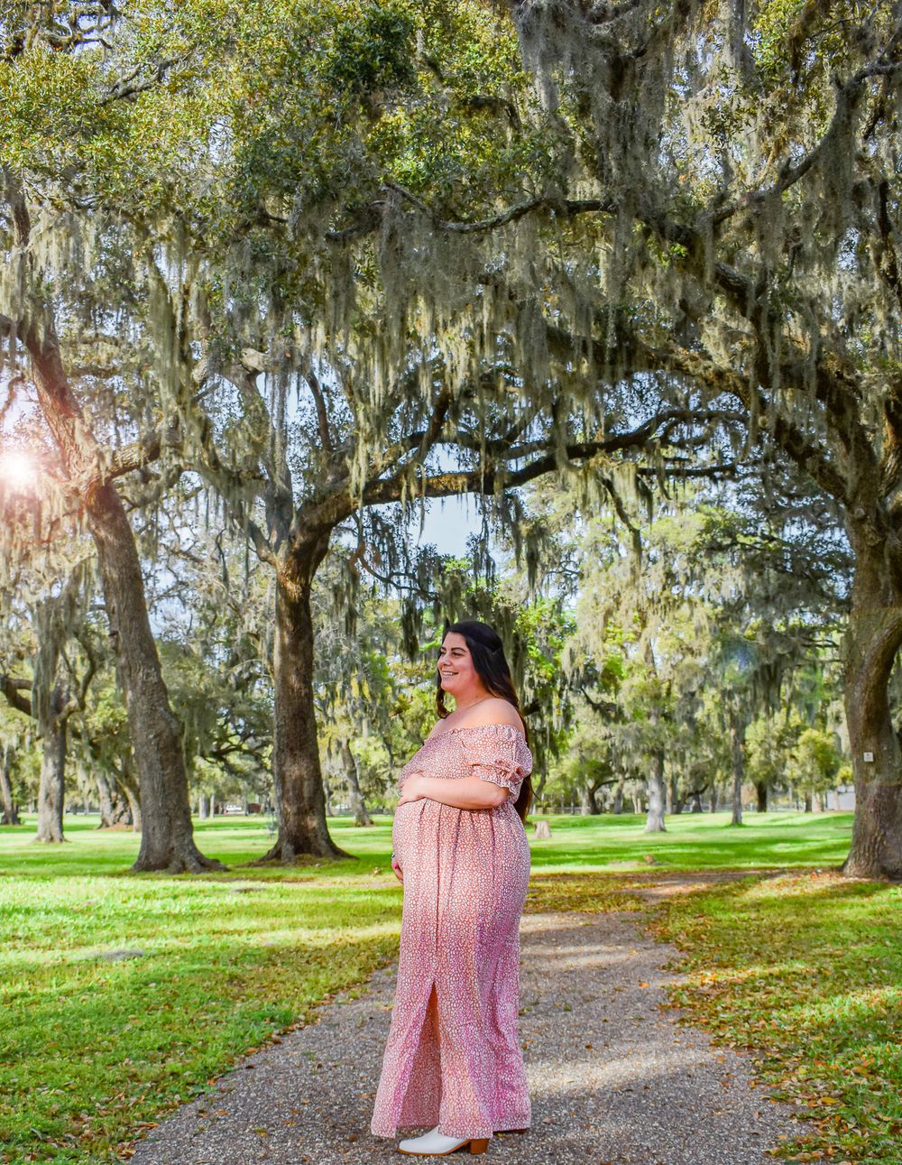 Expectant mother in pink dress standing on path under moss-draped oak trees.
