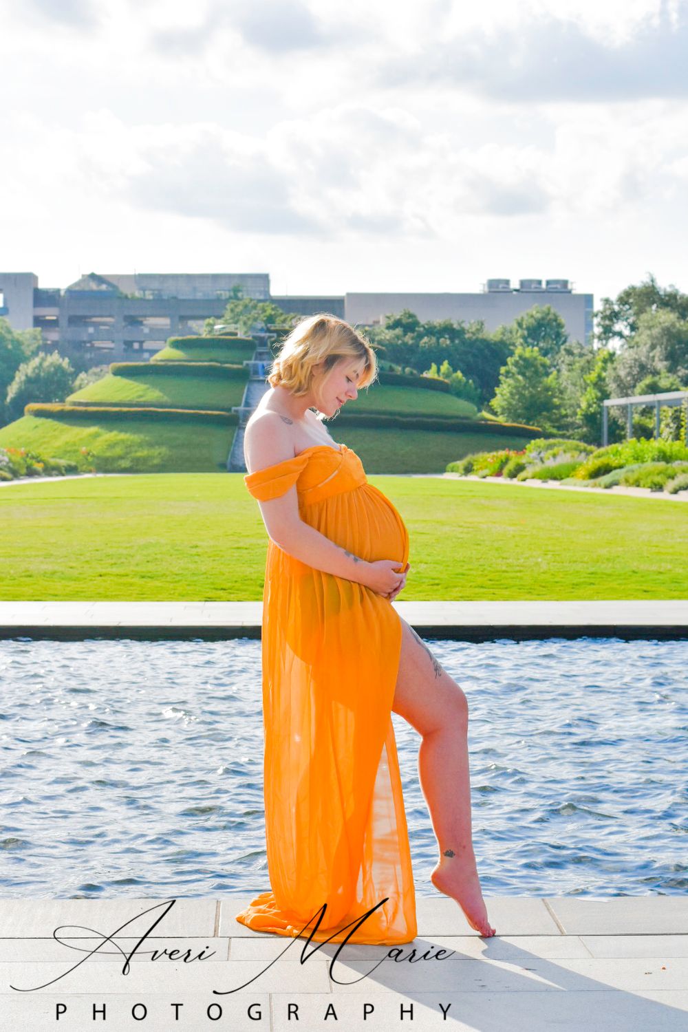 Maternity photo of woman in flowing orange dress standing by pool with scenic landscape.