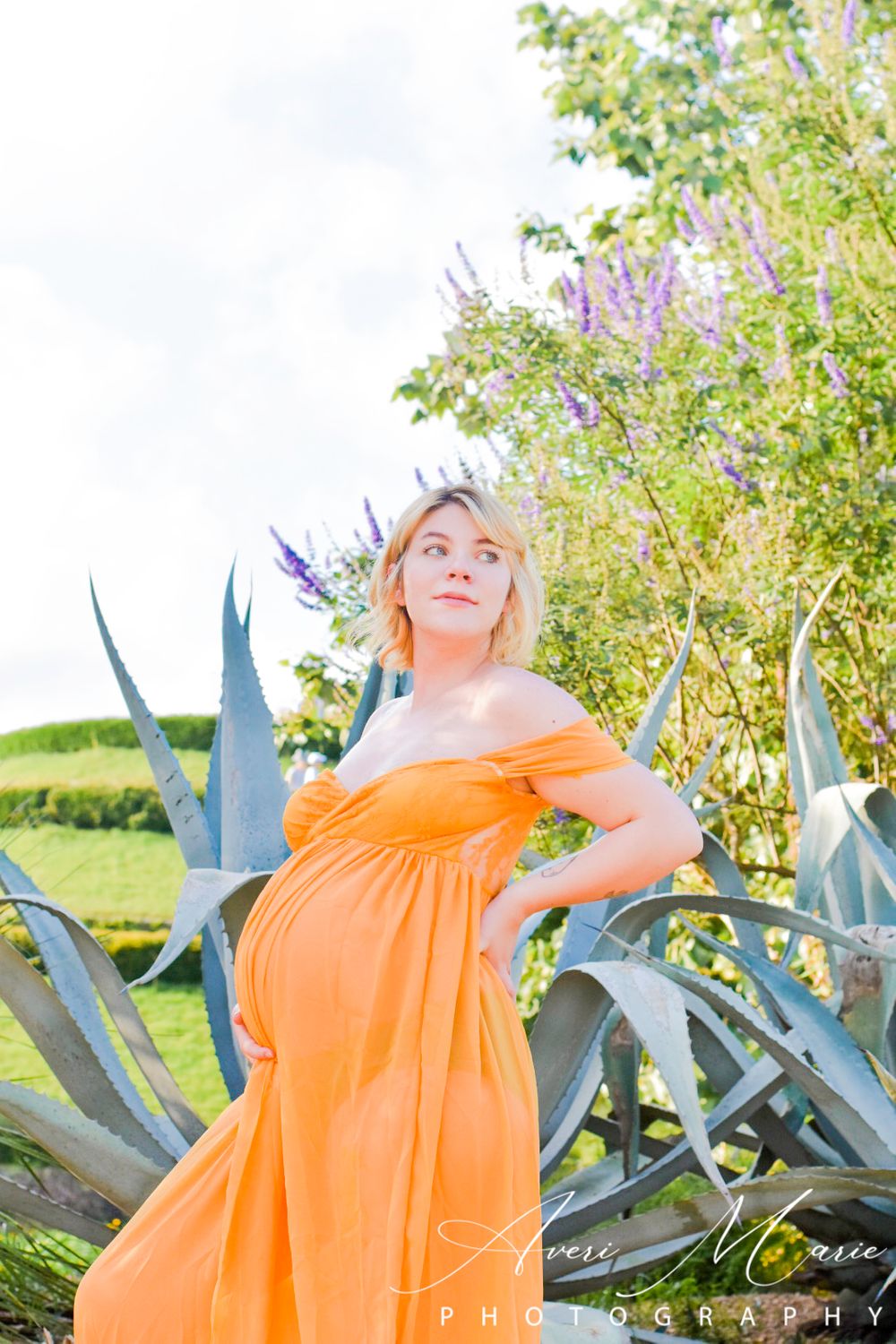 Expectant mother in bright orange dress poses among agave plants in garden setting.