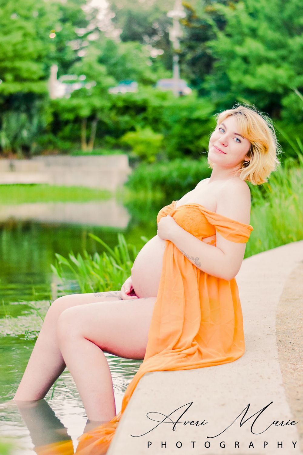 Maternity portrait by a pond with person wearing flowing orange dress.