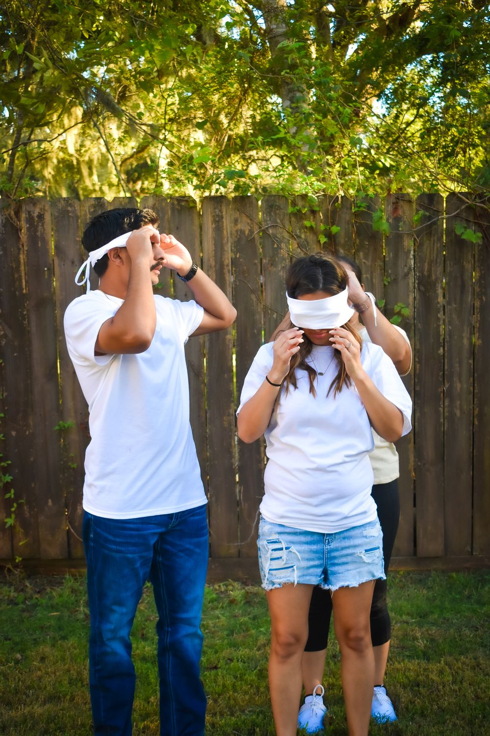 Two people wearing white shirts and blindfolds stand in a backyard with sunlit trees and a wooden fence.