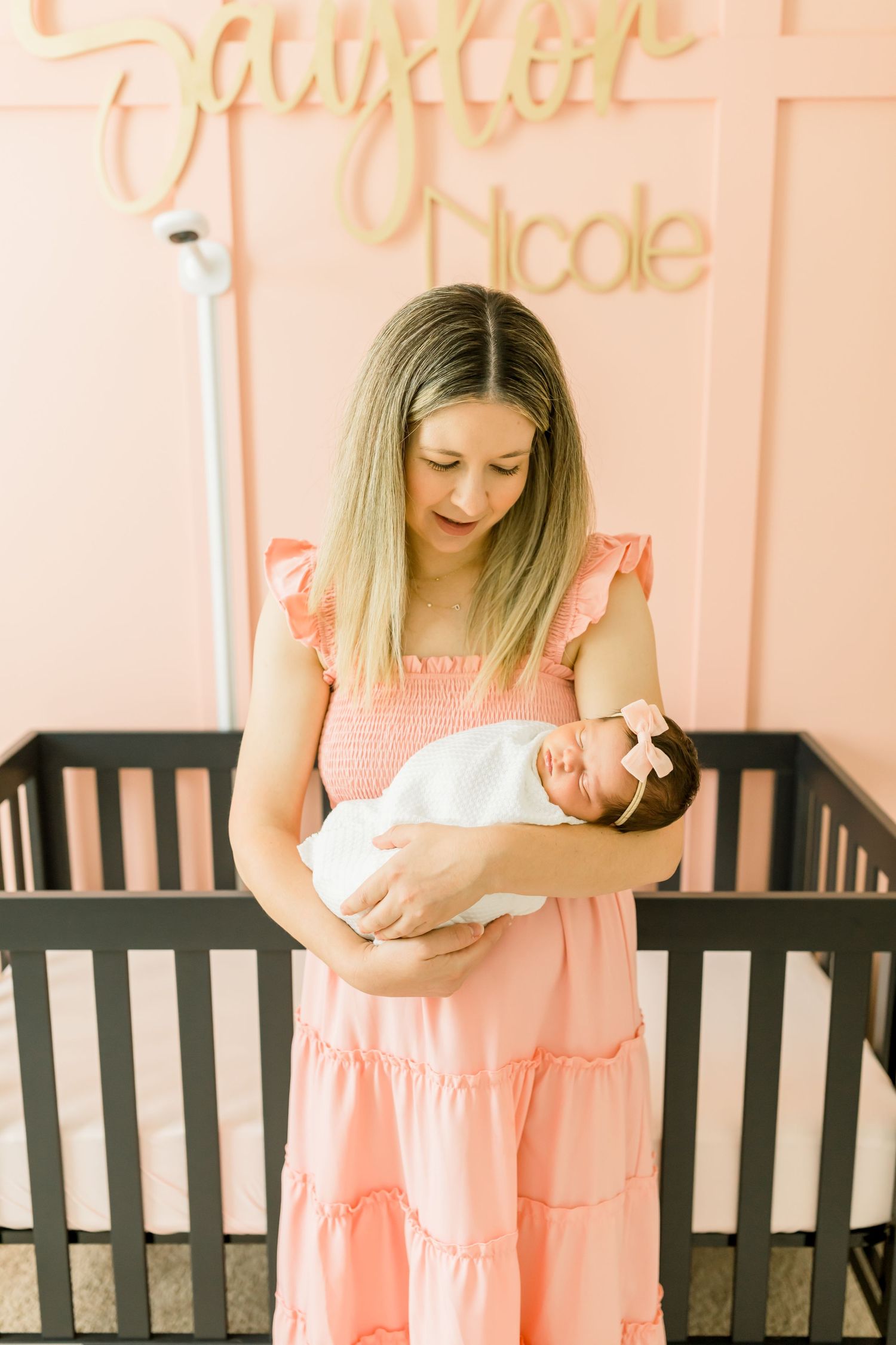 Mother in coral dress holds sleeping newborn by black crib.