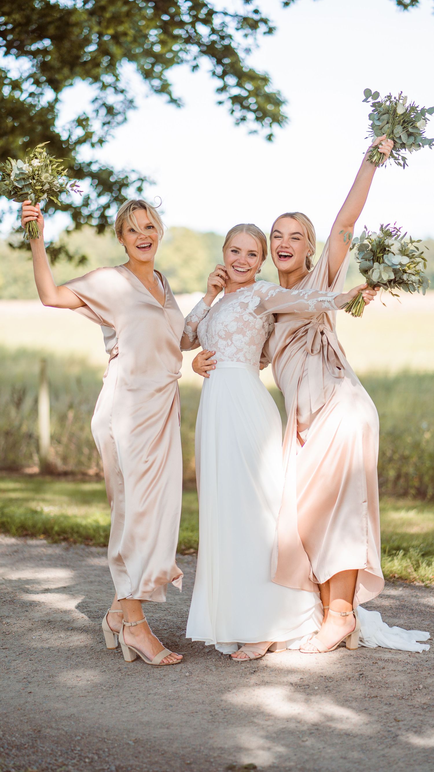 Bride and two bridesmaids in champagne satin dresses cheering with bouquets on sunny path.