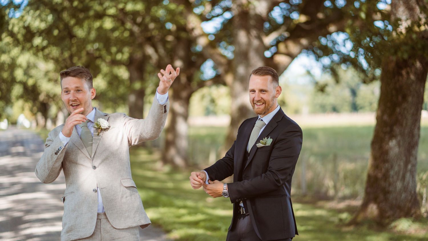 Two groomsmen in suits laughing and dancing playfully on a tree-lined wedding venue path.