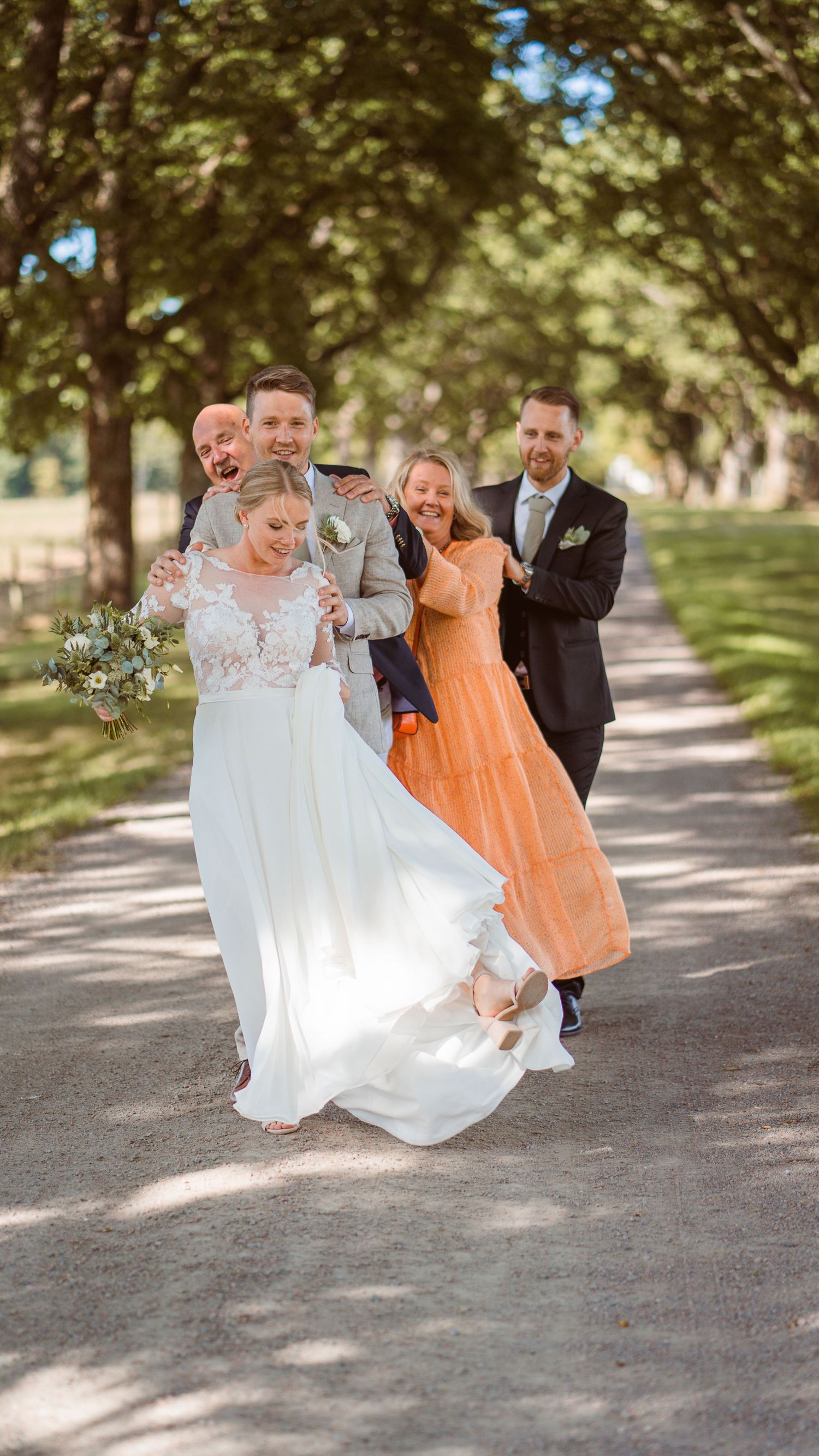 Joyful wedding party celebrating with bride and groom on a sunlit tree-lined avenue.