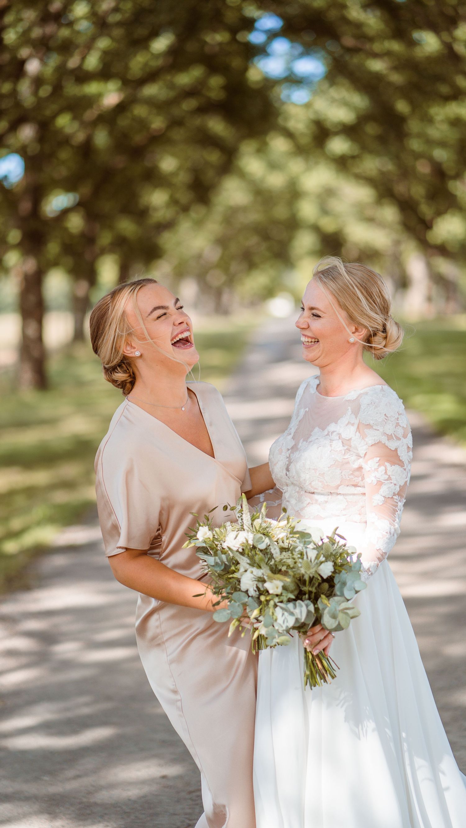Bride in lace gown and bridesmaid in champagne dress laughing joyfully on tree-lined path.