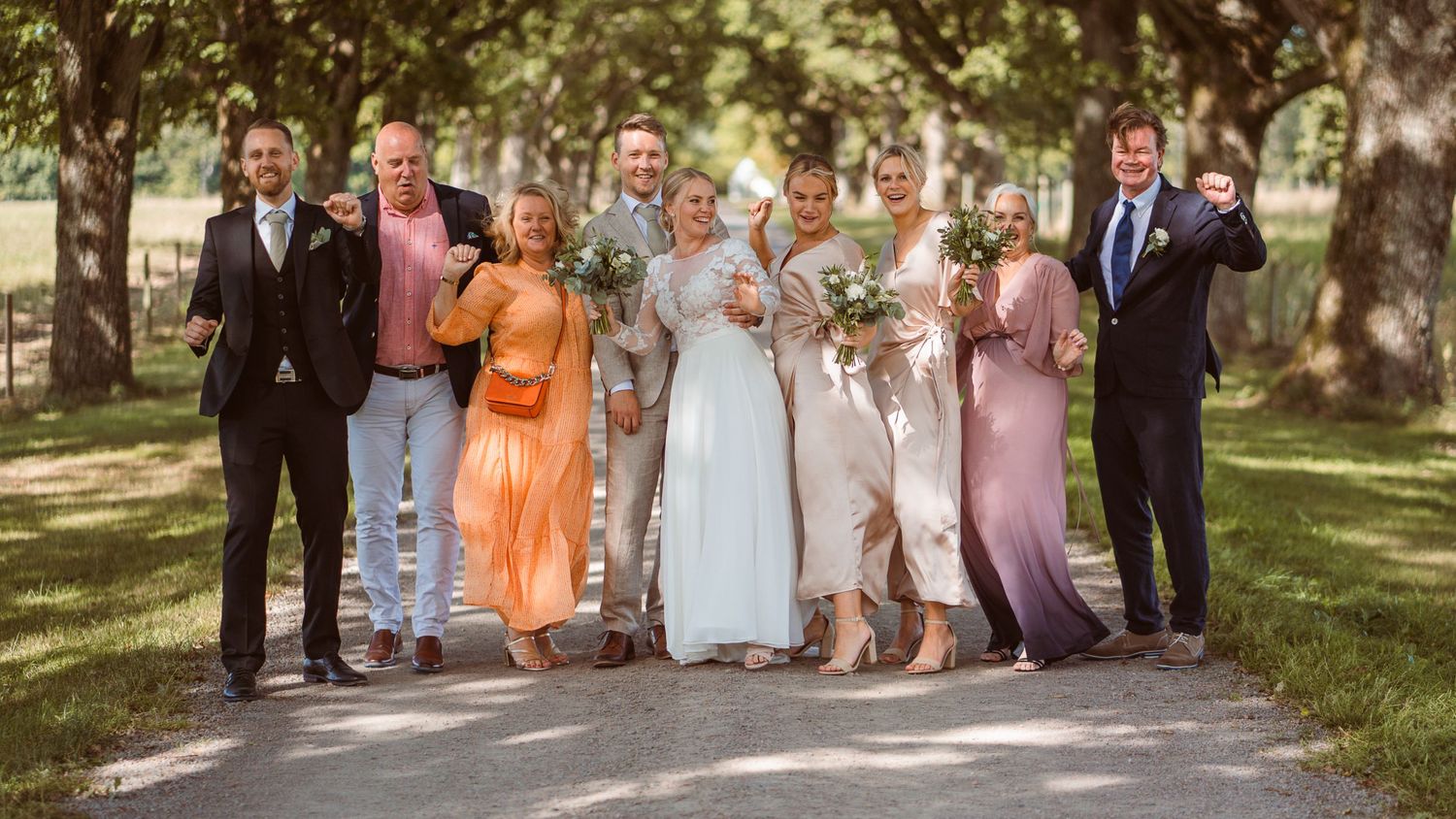 Happy wedding party group photo with bride and groom surrounded by family on tree-lined path.