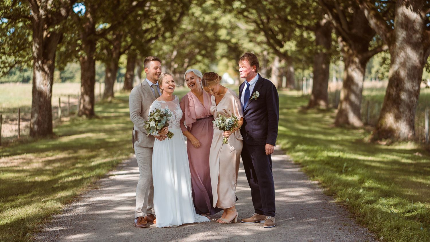 Wedding party of four walking and laughing along a sunny tree-lined path.